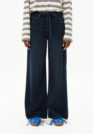 Person trägt dunkle Jeans mit weitem Bein und Bindeband in der Taille, ein gestreiftes Langarmshirt und leuchtend blaue Turnschuhe mit gelben Schnürsenkeln.
