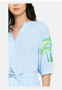 Camicia a righe blu chiaro con bottoni frontali, maniche corte a sbuffo e ricamo di palme verdi sul braccio sinistro. Si annoda in vita.