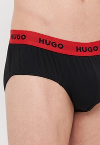 Schwarze Unterhose mit einem roten Gummibund, auf dem "HUGO" in Schwarz steht. Der Stoff wirkt glatt mit einem taillierten Design und normalem Schnitt.
