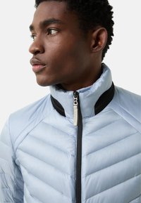 Light blue quilted jacket with a high collar and black accents, featuring a front zipper and textured fabric in a chevron pattern.