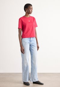 Rød bomulds t-shirt med korte ærmer og rund halsudskæring samt et lille logo. Combineret med lyseblå brede jeans og sorte slip-on sko.