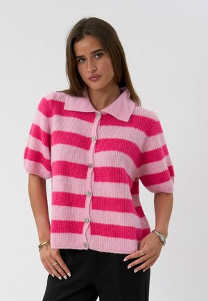 Vrouw draagt een kort mouw, gebreid vest met knopen en brede horizontale strepen in roze en fuchsia, staand met handen bij de heupen tegen een effen achtergrond.