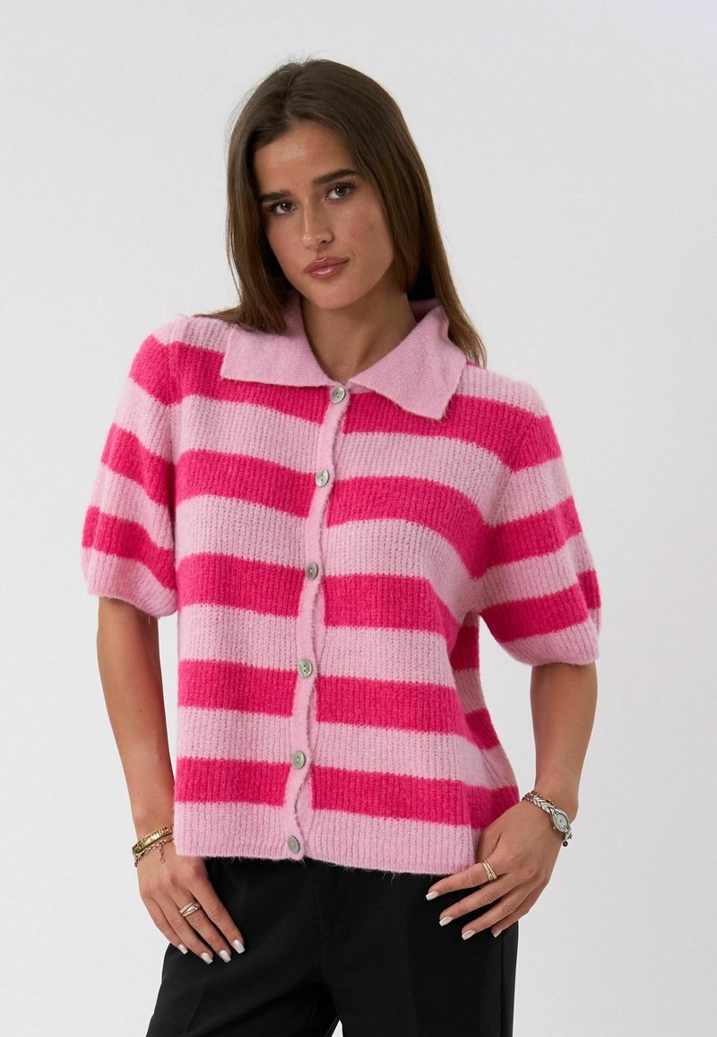 Femme portant un cardigan à manches courtes, boutonné, avec de larges rayures horizontales roses et fuchsia, debout les mains près des hanches, devant un fond uni.