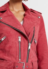 Czerwona zamszowa kurtka motocyklowa z srebrnymi zamkami, dużym kołnierzem i paskiem w talii. Posiada kieszenie na zamek i teksturowane wykończenie.