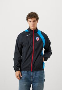 Nike vindjakke i sort med blå detaljer, rød lynlås og Atlético Madrid-logo. Den har høj krave og elastiske mansjetter.