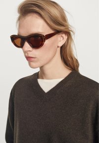 Mladá osoba se světlými hnědými vlasy, která má na sobě hnědé želvovinové brýle a tmavě hnědý svetr s výstřihem do V přes bílou košili.