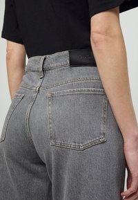 Person, der hochgeschnittene graue Denim-Jeans mit Gesäßtaschen und einem schwarzen Lederetikett am Bund trägt, kombiniert mit einem schwarzen Kurzarmoberteil.