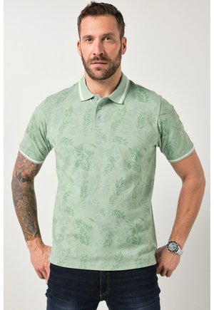 Homme portant un polo vert clair à motif de feuilles, un jean foncé, une montre au poignet gauche et un tatouage sur l'avant-bras droit, debout face à l'avant.