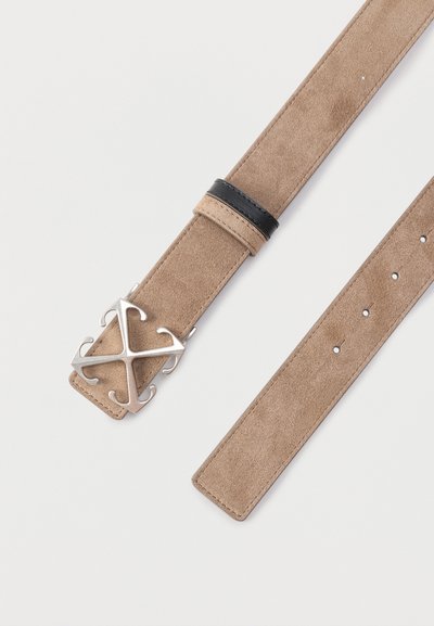 Ζώνη από σουέντ ανοιχτού καφέ χρώματος με πολλαπλές τρύπες και ασημί μεταλλική αγκράφα σε σχήμα στυλιζαρισμένου σταυρού, με μαύρο δερμάτινο θηλάκι.