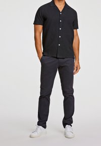 Chemise noire à manches courtes avec boutons, associée à un pantalon bleu marine et des baskets blanches. La chemise a un col et des boutons blancs. Coupe classique.