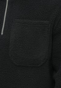 Pulover negru din flece, cu un design cu fermoar pe un sfert, având o suprafață texturată și un buzunar mare în stil patch pe față.