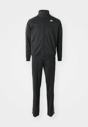 Tuta nera realizzata in poliesteri lucido. Presenta una giacca con zip e colletto alto e pantaloni abbinati con strisce laterali e logo Kappa.
