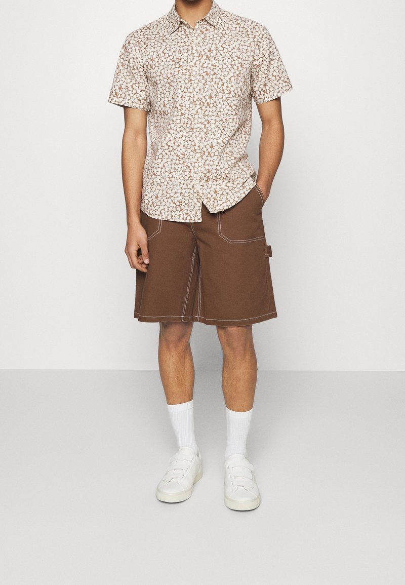 Homme portant une chemise à fleurs à manches courtes, un short marron longueur genou avec des coutures blanches, des chaussettes blanches et des baskets blanches, debout à l'intérieur.