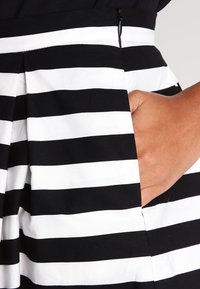 Ruka odpočívající uvnitř kapsy černobílé sukně s horizontálními pruhy a zipovým detailem v pase.