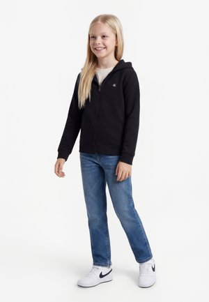 Fată zâmbitoare cu păr blond lung, purtând un hanorac negru cu fermoar, blugi albaștri și adidași albi, stând în fața unui fundal alb.