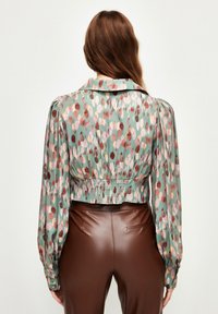 Blusa corta in tessuto verde a fantasia con pois rosa e marroni, con maniche a palloncino e schiena arricciata, abbinata a pantaloni marroni aderenti.