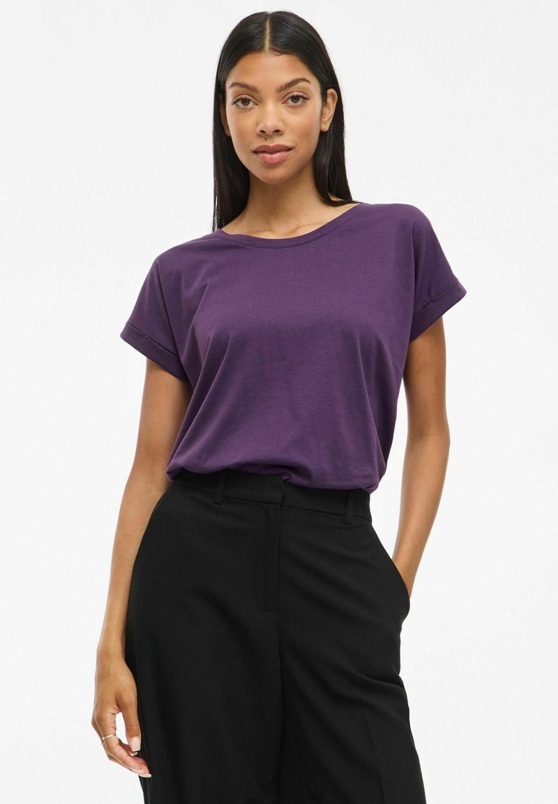 Kvinna med långt mörkt hår som bär en lila kortärmad skjorta instoppad i högmidjade svarta byxor med händerna i fickorna, neutral bakgrund.