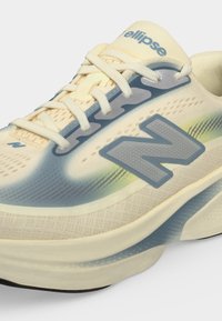 Ανοιχτό μπεζ και μπλε αθλητικό παπούτσι New Balance με κορδόνια, υφή στη σόλα και λογότυπο "N" στο πλάι, όνομα μοντέλου "ellipse" στη γλώσσα.