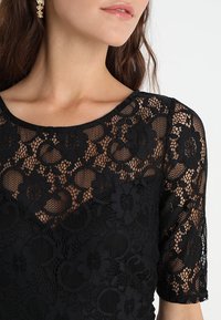 Top en dentelle noire avec un motif floral, des manches courtes et un col rond, révélant une texture transparente sur la peau.