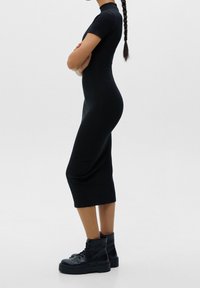 Femme debout, les bras croisés, portant une robe midi noire ajustée et des bottines noires lacées, avec une longue queue de cheval tressée.