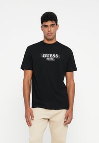 Camiseta de algodón negra con mangas cortas que presenta el logo blanco "GUESS" y un gráfico del año de establecimiento en el frente. Diseño simple, corte relajado.