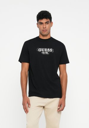 Camiseta de algodón negra con mangas cortas que presenta el logo blanco "GUESS" y un gráfico del año de establecimiento en el frente. Diseño simple, corte relajado.