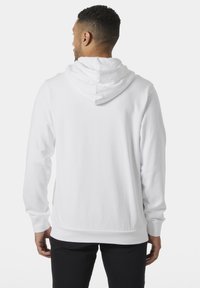 Hombre con sudadera con capucha blanca lisa visto desde atrás, de pie contra un fondo claro.