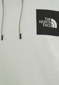 Sudadera con capucha de color gris claro que presenta un logo cuadrado negro con el texto "THE NORTH FACE". Incluye cordones con puntas negras y una superficie suave y texturizada.