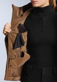 Smeđi izolirani sako s zatvaračem sprijeda, ispod kojeg se nalazi crni rebrasti pulover s visokim ovratnikom i džepom koji drži dodatke.