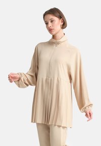 Top beige con zip e colletto alto, con maniche elastiche, caratterizzato da un design plissettato in vita, realizzato in un tessuto morbido e leggero.
