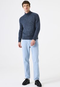Man die staat met een donkerblauwe gebreide trui, lichtblauwe jeans en zwarte veterschoenen voor een effen witte achtergrond.