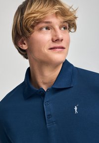 Navyblaues Poloshirt aus einem strukturierten Stoff, mit einem Drei-Knopf-Verschluss und kleinem weißen Logo auf der linken Brust. Kurzärmlig und mit klassischem Kragendesign.