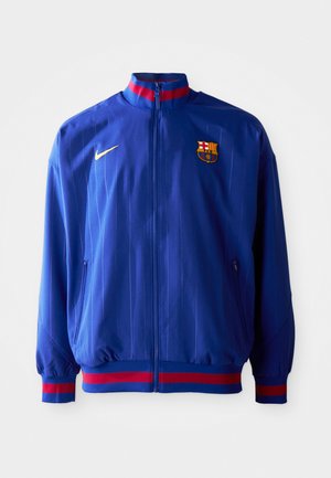 Plava Nike trenirka s vertikalnim prugama, visokim ovratnikom i crvenim i plavim detaljima. Uključuje džepove sa patentnim zatvaračem i logo kluba.