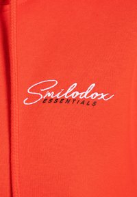 Sweatshirt orange vif avec une texture douce, présentant un logo brodé blanc "Smilodox ESSENTIALS" et un cordon de serrage tressé.