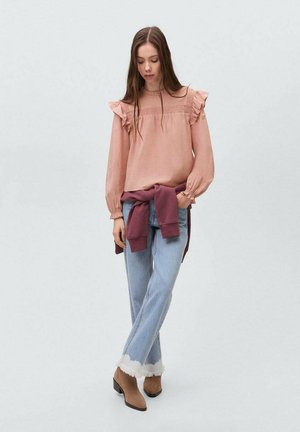Lichtroze blouse met ruches bij de schouders en lange mouwen, gecombineerd met lichtblauwe jeans met rafelige zomen en een bordeauxrode sweatshirt geknoopt om de taille.