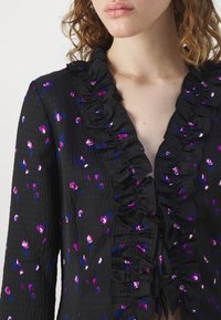 Gros plan d'une femme portant un chemisier noir texturé avec un imprimé floral violet et bleu et des volants le long de l'encolure et à l'avant.