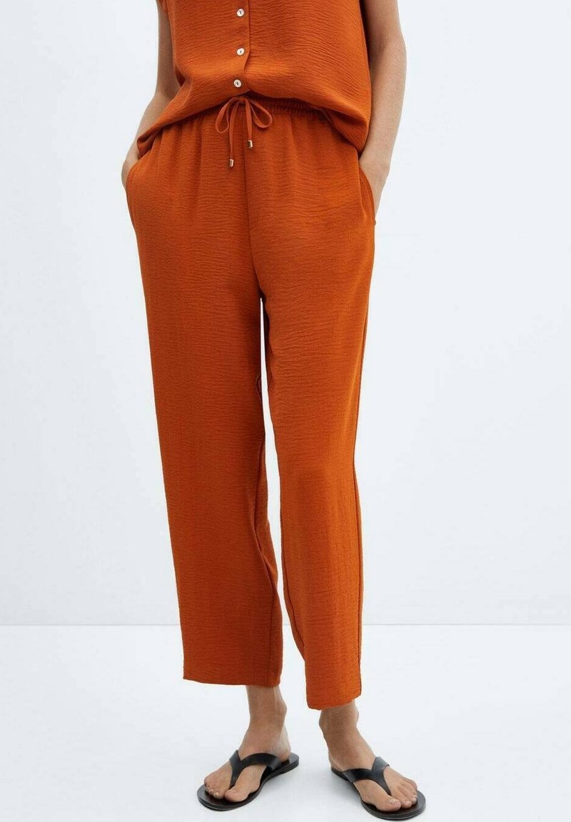 Femme portant un pantalon ample couleur orange brûlé avec une taille à cordon et des sandales noires, debout les mains dans les poches.