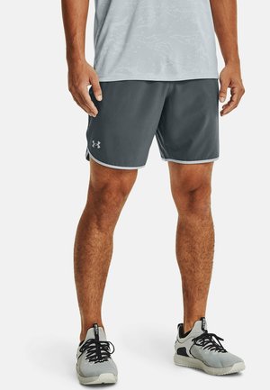 Mann trägt graue Sportshorts mit Under Armour-Logo und graue Laufschuhe, steht vor weißem Hintergrund.