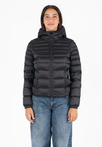 Zwarte, pufferjack met rits en capuchon, met horizontale stiknaden en elastische manchetten. Draagt over lichtblauwe, losse jeans.