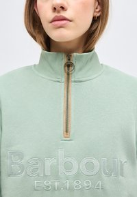 Νεαρή γυναίκα που φοράει ανοιχτό πράσινο φούτερ Barbour με φερμουάρ στο μισό και ψηλό γιακά, με ορατό κέντημα "Barbour EST. 1894" στο μπροστινό μέρος.