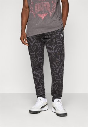 Mand iført sorte mønstrede Puma sweatpants, hvide sportssko og en grå ærmeløs grafisk t-shirt, der står foran en ensfarvet baggrund.