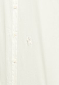 Camisa de cor creme feita de tecido macio, com fecho de botões na frente e um pequeno logótipo AX bordado na lateral.