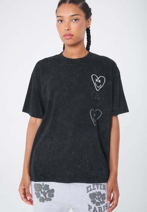 Joven mujer con el cabello trenzado que lleva una camiseta negra oversize con diseños de corazones y pantalones cortos grises con patrones florales y de texto.
