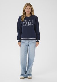 Navyblauwe sweater met witte tekst "C'est la vie PARIS" op de borst. Gecombineerd met lichtblauwe wijde jeans en beige sneakers.