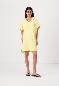 Gul frottéklæde poncho med hætte og korte ærmer, forlommer og dekorativt sort kantbånd i kanten. Stilfuld og afslappet design.