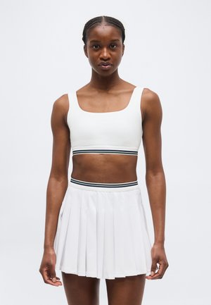 Γυναίκα με πλεξούδες, που φοράει άσπρο crop top και πλισέ φούστα με μαύρες ρίγες στη μέση, στέκεται μπροστά σε ένα απλό λευκό φόντο.