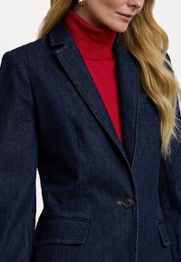 Temno modri blazer iz denima z enim gumbom, zareznimi ovratniki in dvema sprednjima žepoma. Nosi se čez rdeč pulover s stojalom. Gladka tekstura in prilagojen kroj.