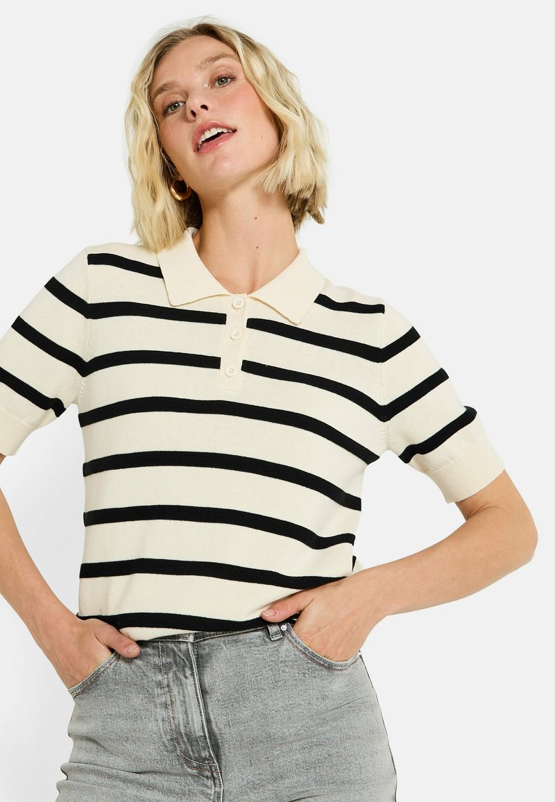 Mujer rubia con cabello corto, vistiendo una camiseta de polo a rayas crema y negra y jeans gris claro, con las manos en los bolsillos, de frente.