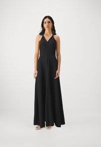 Rochie maxi neagră, fără mâneci, cu gât V, fustă evazată și textură fină. Pur rită cu sandale de culoare deschisă și cercei aurii.
