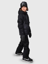 Niño con ropa de invierno negra que lleva gafas de esquí, manoplas y pantalones de nieve, de pie contra un fondo simple.